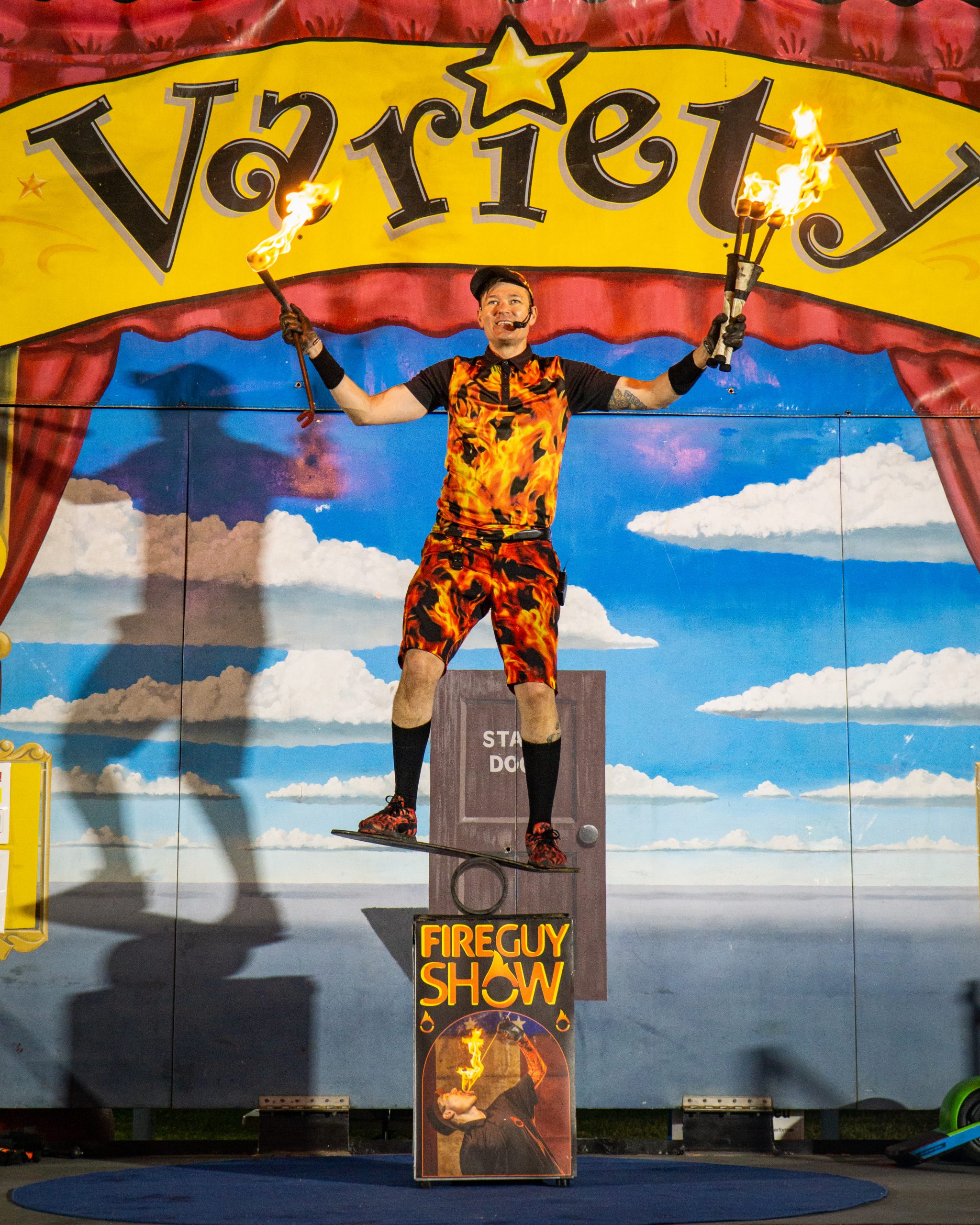 FireGuy is balancing on a plank, on a pipe, on top of a box inside on a stage, while juggling four fire sticks.