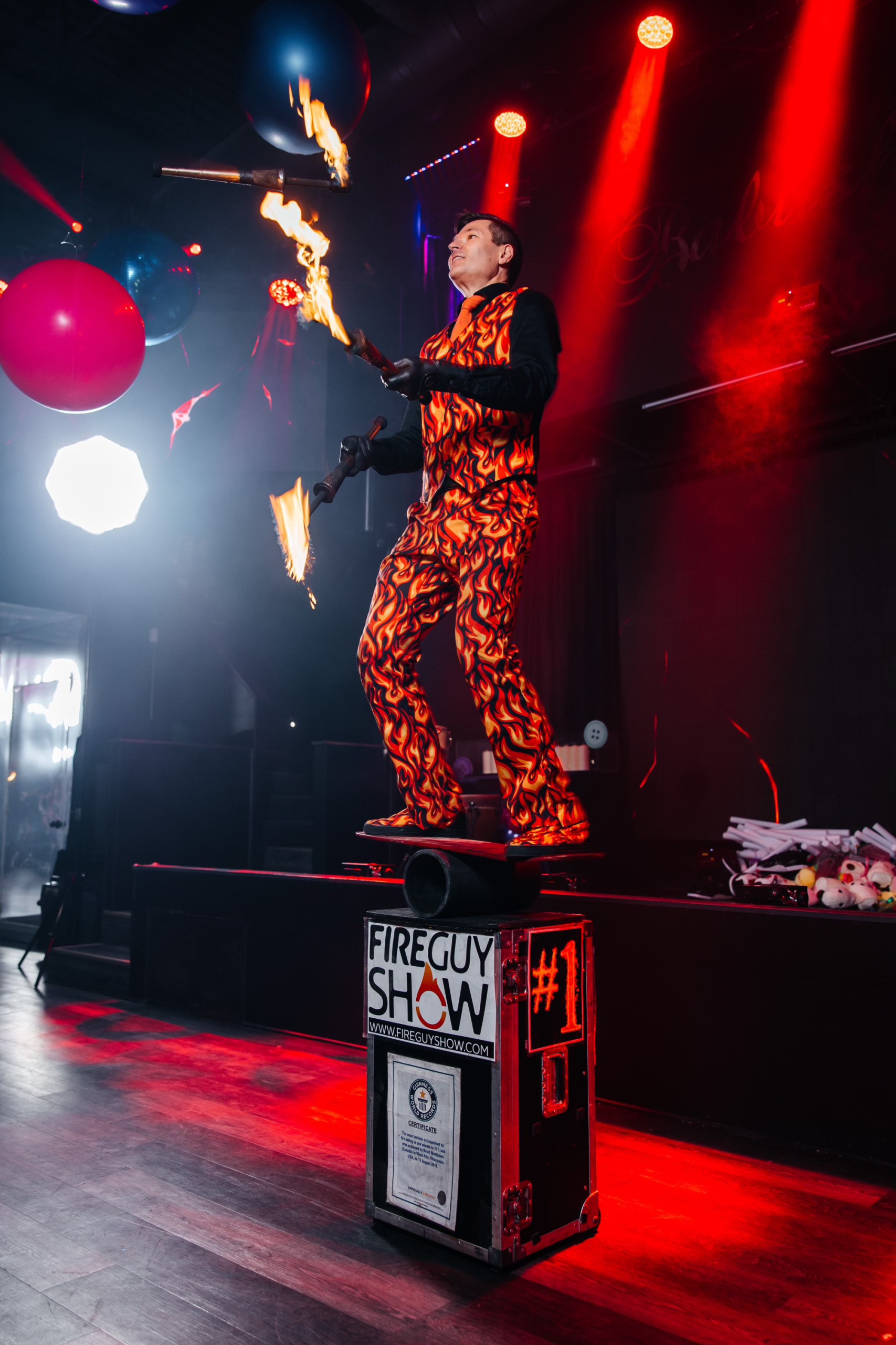 FireGuy standing on a board, on a pipe, on a box and juggling while balancing.